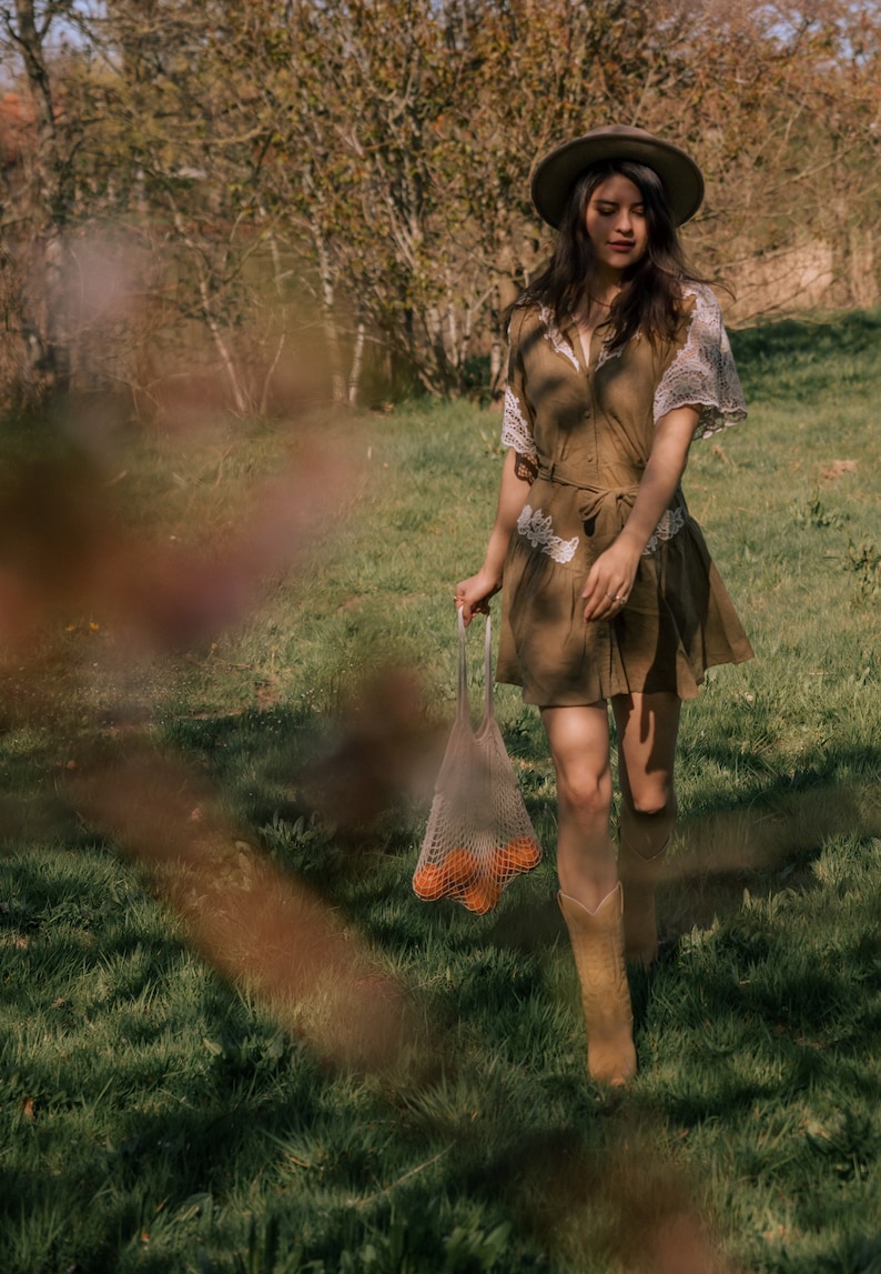 Puede incluir: Una persona con un vestido marr&oacute;n, sombrero y botas camina por un campo cubierto de hierba, sosteniendo una bolsa de malla llena de naranjas. El vestido tiene mangas de encaje y un cintur&oacute;n. El sombrero es verde oliva.