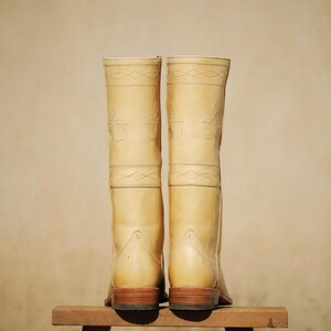 May include: A pair of light tan leather cowboy boots with decorative stitching. The boots are tall, with a classic design, and sit on a small wooden stool. The soles are a dark brown color.