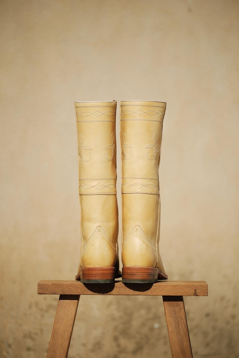 May include: A pair of light tan leather cowboy boots with decorative stitching. The boots are tall, with a classic design, and sit on a small wooden stool. The soles are a dark brown color.