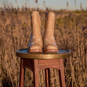 May include: A pair of tan leather ankle boots with white daisy embroidery. The boots are sitting on a wooden stool with a brass top.