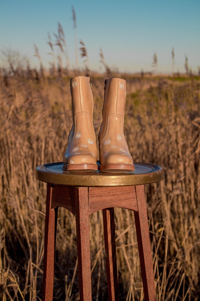 May include: A pair of tan leather ankle boots with white daisy embroidery. The boots are sitting on a wooden stool with a brass top.