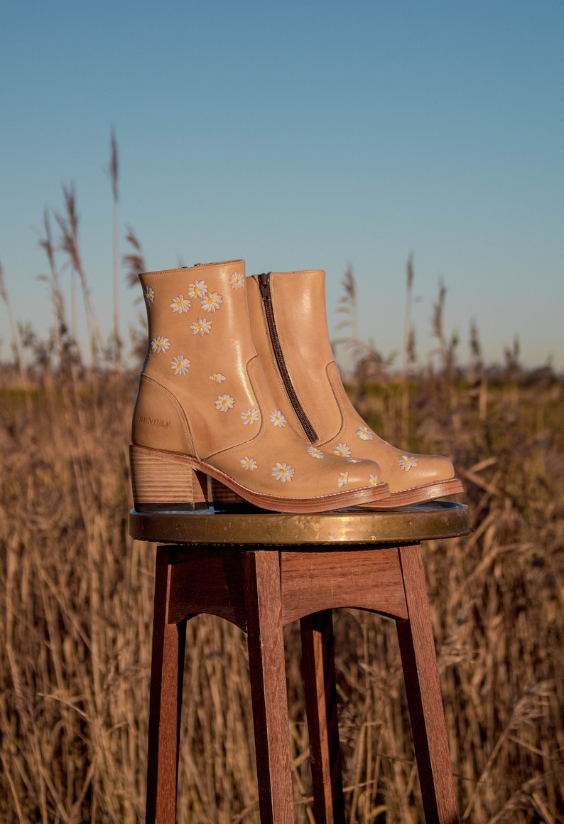 May include: A pair of tan leather ankle boots with a daisy print. The boots have a block heel and a zipper on the side. The boots are sitting on a wooden stool.