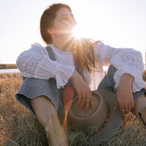 May include: A woman wearing a white lace top and denim overalls sits in a field of tall grass, holding a brown felt hat. The sun shines brightly behind her, creating a warm glow.
