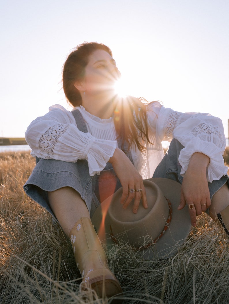 May include: A woman wearing a white lace top and denim overalls sits in a field of tall grass, holding a brown felt hat. The sun shines brightly behind her, creating a warm glow.
