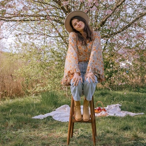 May include: A woman wearing a floral print blouse, blue jeans, and brown boots sits on a wooden stool in a grassy field. A cherry blossom tree is in the background.