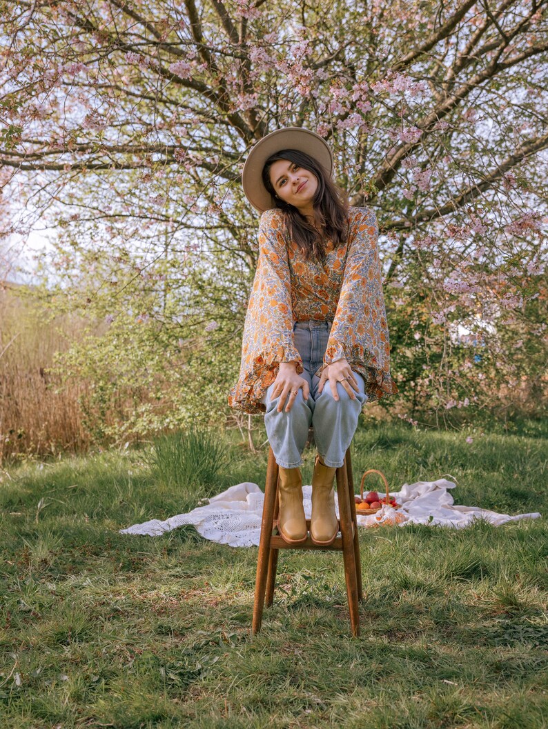 May include: A woman wearing a floral print blouse, blue jeans, and brown boots sits on a wooden stool in a grassy field. A cherry blossom tree is in the background.