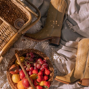 Puede incluir: Una escena de picnic con botas de vaquero de cuero color canela, una cesta de mimbre llena de fruta y una silla de mimbre. La cesta contiene manzanas rojas, fresas, uvas y limones. Las botas tienen punta cuadrada y tac&oacute;n apilado.