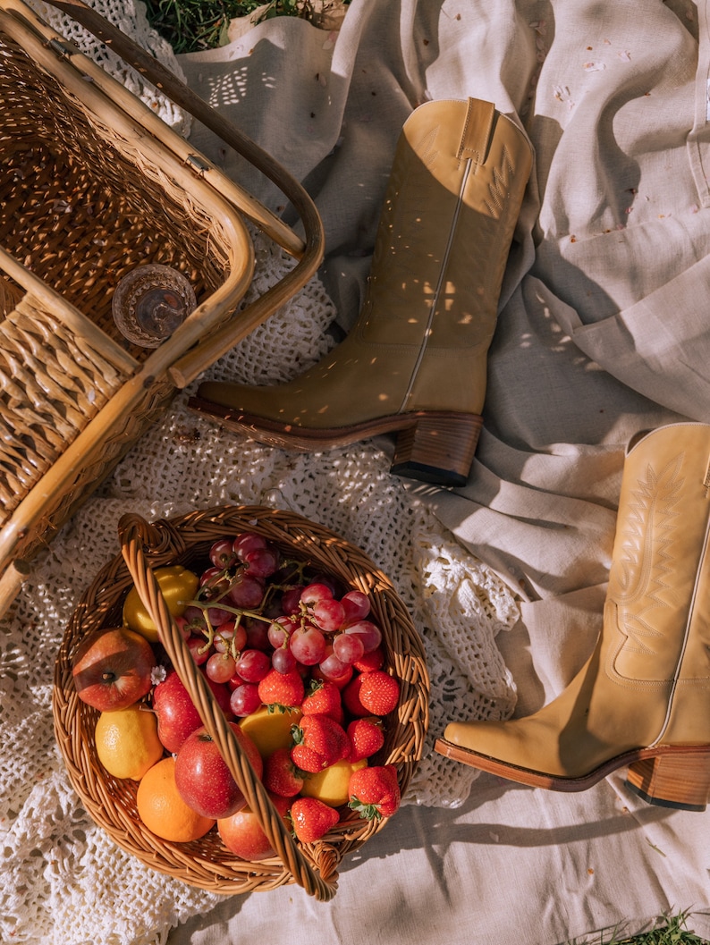 Puede incluir: Una escena de picnic con botas de vaquero de cuero color canela, una cesta de mimbre llena de fruta y una silla de mimbre. La cesta contiene manzanas rojas, fresas, uvas y limones. Las botas tienen punta cuadrada y tac&oacute;n apilado.