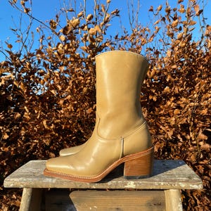 May include: Tan leather cowboy boots with a block heel. The boots are sitting on a wooden stool against a backdrop of brown branches and a blue sky.