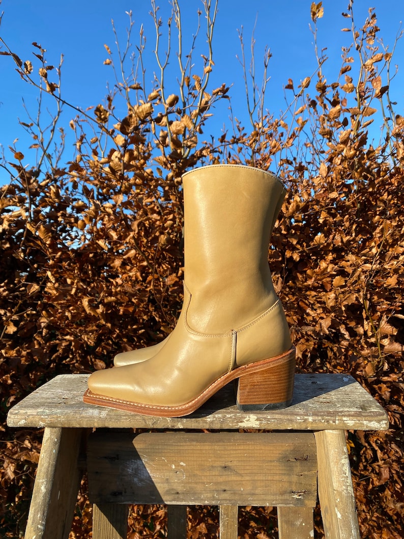 May include: Tan leather cowboy boots with a block heel. The boots are sitting on a wooden stool against a backdrop of brown branches and a blue sky.