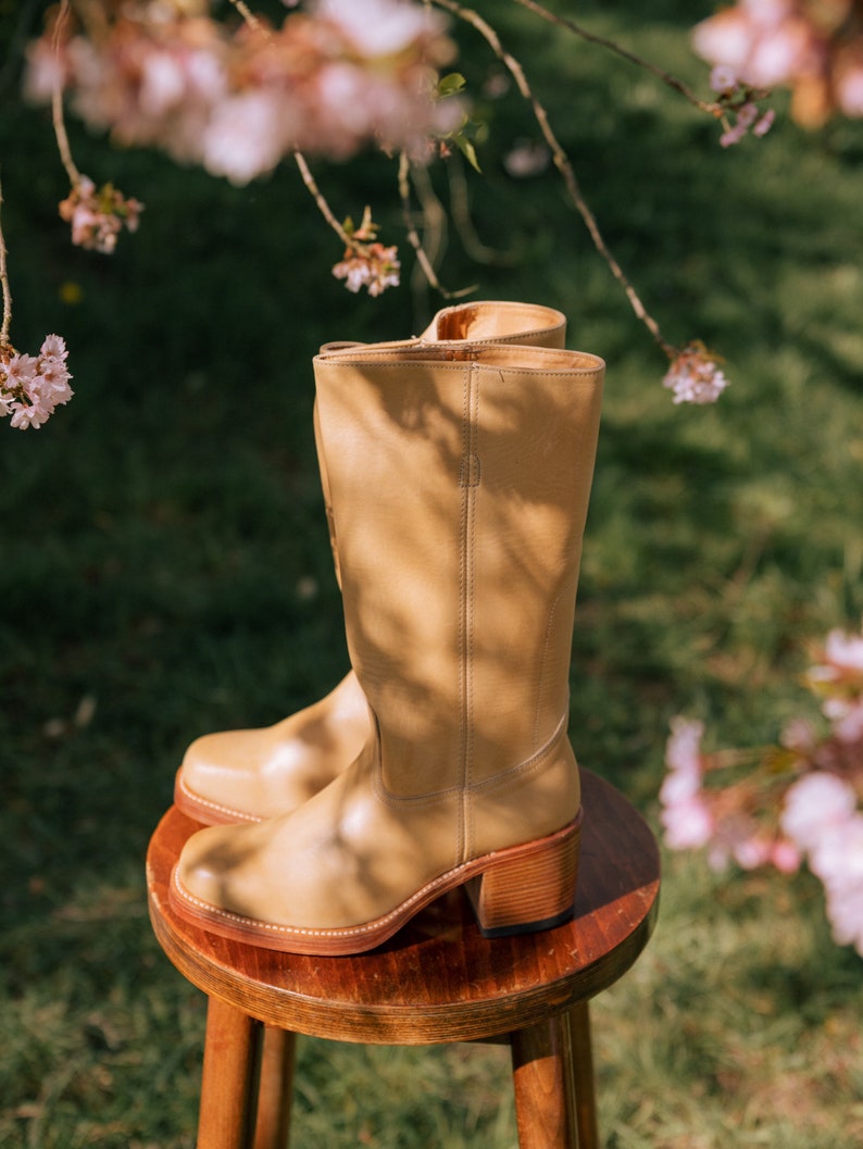 May include: A pair of tan leather boots with a stacked wooden heel, resting on a small wooden stool. The boots have a classic design with a rounded toe and a mid-calf height. The background features green grass and pink blossoms.