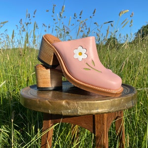 Op de afbeelding: Roze leren sandalen met hak en een wit madeliefje ontwerp. De sandalen staan op een houten kruk met een messing bovenblad.
