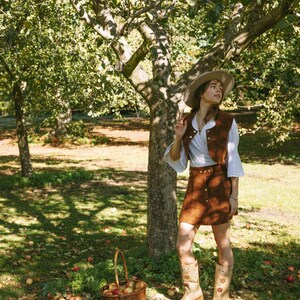 May include: A woman wearing a brown suede skirt and vest, a white blouse, and a straw hat stands in an apple orchard. She is holding a basket of apples and looking up at the trees.