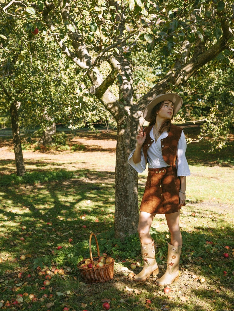 May include: A woman wearing a brown suede skirt and vest, a white blouse, and a straw hat stands in an apple orchard. She is holding a basket of apples and looking up at the trees.