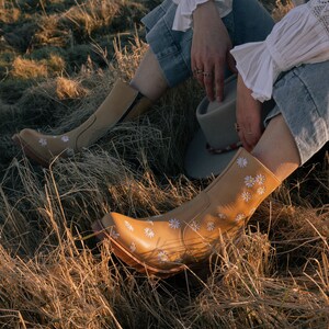 May include: A pair of tan leather ankle boots with white daisy embroidery. The boots are worn by a person sitting in a field of dry grass.