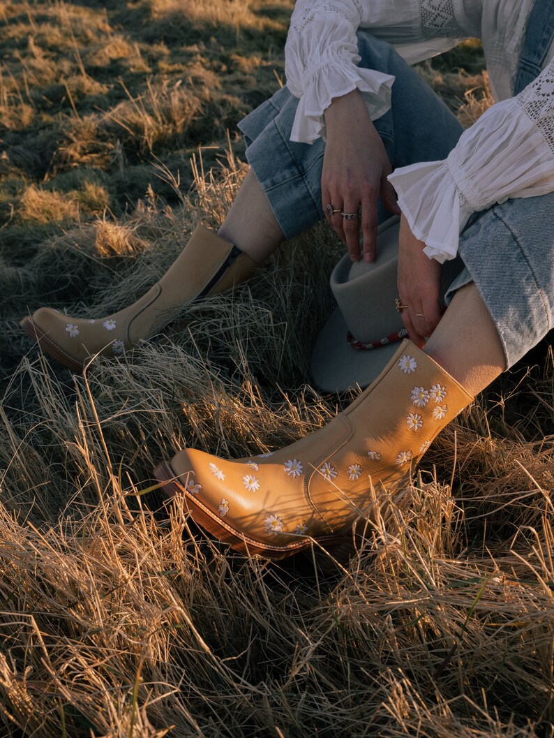 May include: A pair of tan leather ankle boots with white daisy embroidery. The boots are worn by a person sitting in a field of dry grass.