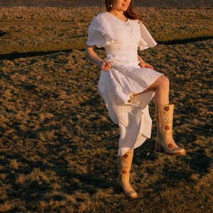 May include: A woman wearing a white dress and a beige hat stands in a field of brown grass. She is wearing beige boots with floral embroidery. The sun is shining brightly.