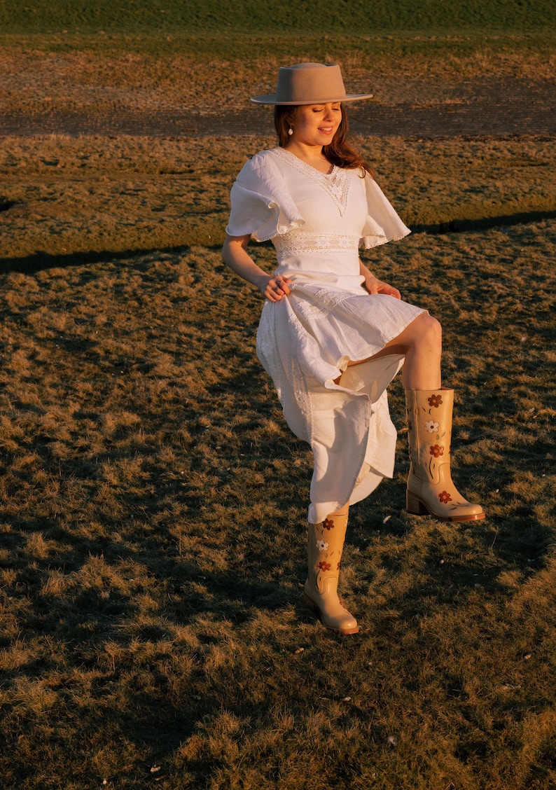 May include: A woman wearing a white dress and a beige hat stands in a field of brown grass. She is wearing beige boots with floral embroidery. The sun is shining brightly.