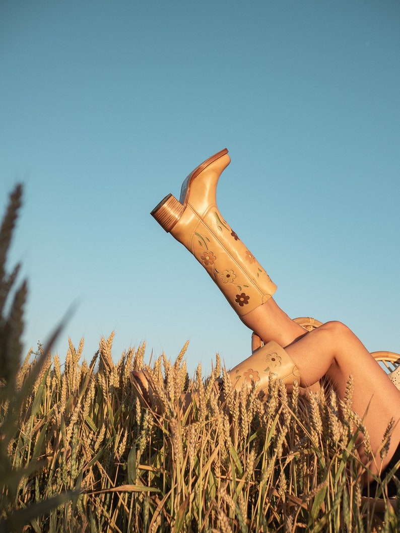 May include: A pair of tan leather boots with floral embroidery, worn by a person lying in a field of wheat.