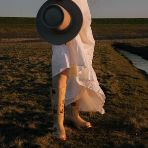 May include: A woman wearing a white dress and floral embroidered boots holds a wide-brimmed hat in front of her. She is standing in a grassy field with a stream in the background.