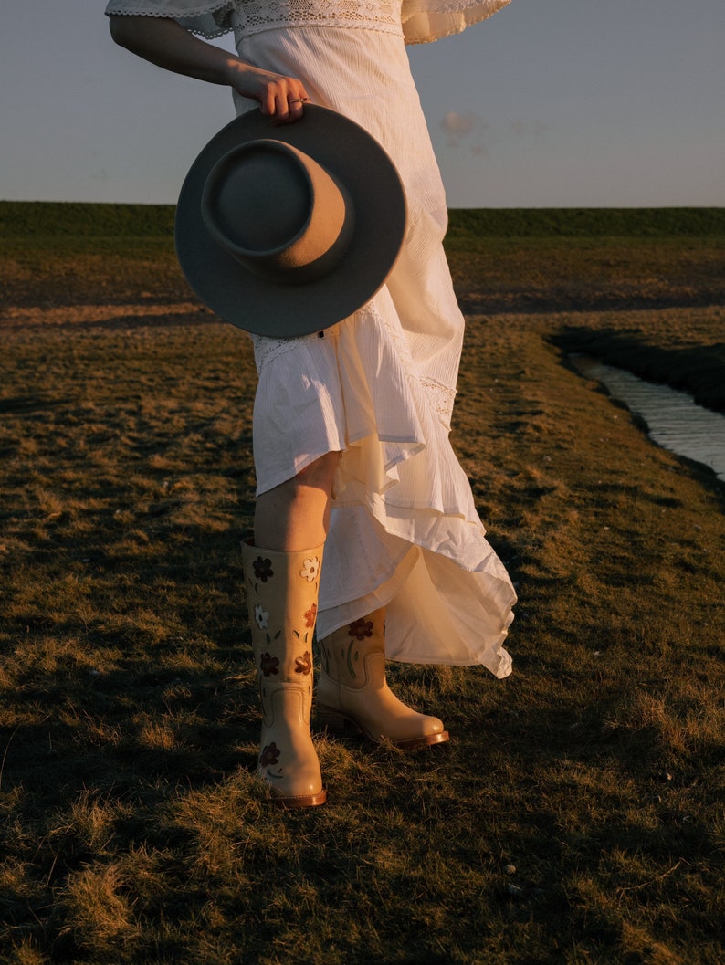 May include: A woman wearing a white dress and floral embroidered boots holds a wide-brimmed hat in front of her. She is standing in a grassy field with a stream in the background.