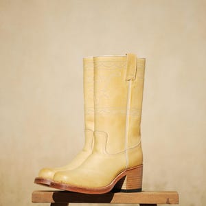 Puede incluir: Un par de botas de vaquero de color amarillo claro con tac&oacute;n grueso y costuras decorativas. Las botas son de cuero y se exhiben sobre un peque&ntilde;o taburete de madera. Las botas tienen un dise&ntilde;o occidental cl&aacute;sico.