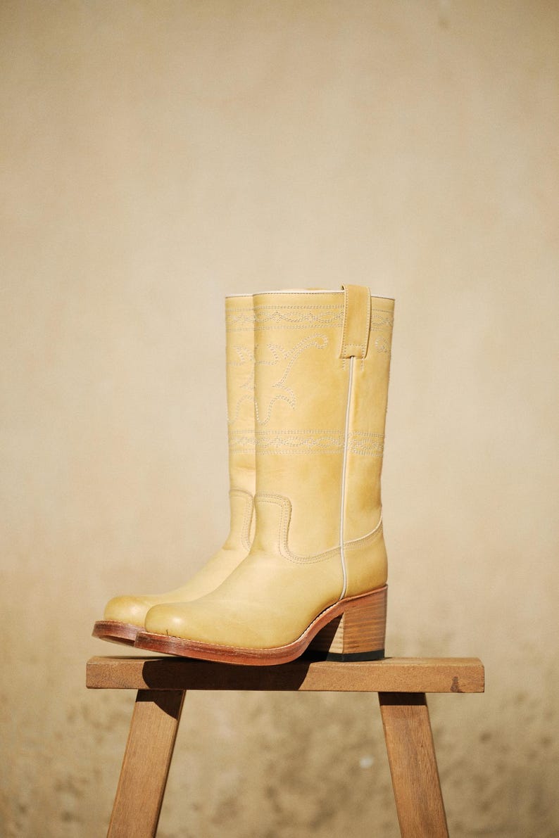 Puede incluir: Un par de botas de vaquero de color amarillo claro con tac&oacute;n grueso y costuras decorativas. Las botas son de cuero y se exhiben sobre un peque&ntilde;o taburete de madera. Las botas tienen un dise&ntilde;o occidental cl&aacute;sico.