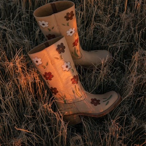 May include: A pair of tan leather cowboy boots with floral embroidery. The boots have a pointed toe and a low heel. The flowers are in shades of red, white, and brown.