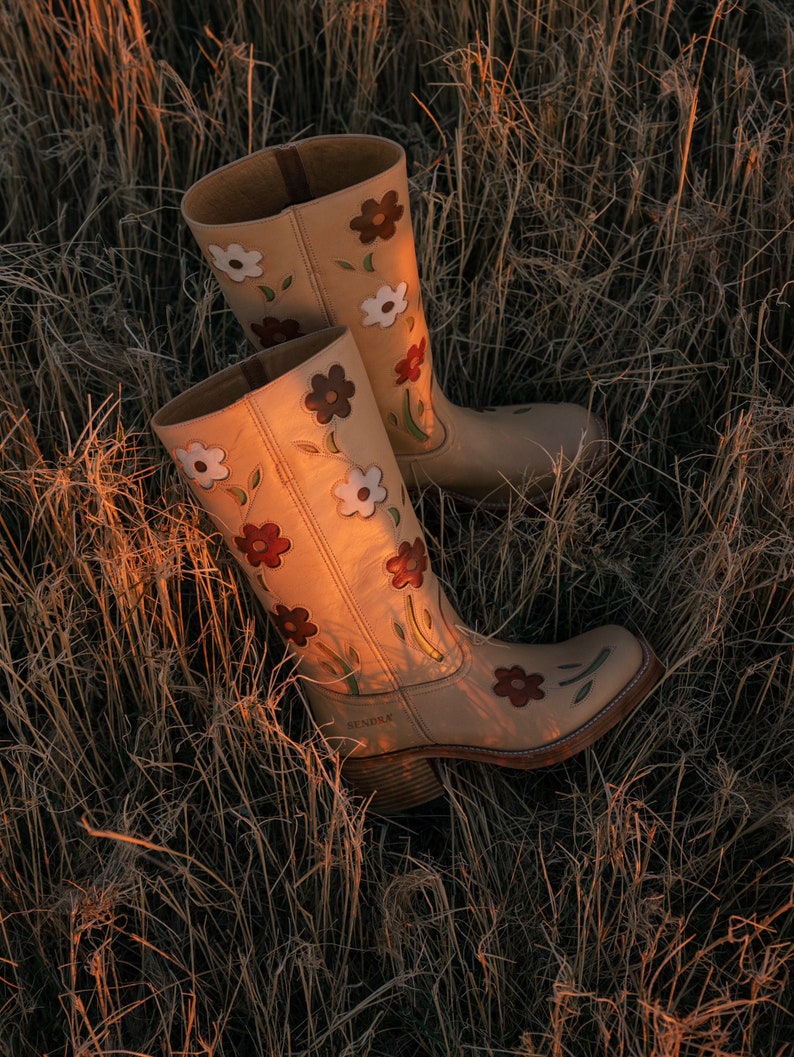 May include: A pair of tan leather cowboy boots with floral embroidery. The boots have a pointed toe and a low heel. The flowers are in shades of red, white, and brown.