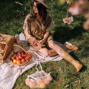 Puede incluir: Una escena de picnic con una mujer con un vestido y sombrero marrones, sentada en una manta en una zona de c&eacute;sped. Una cesta de mimbre llena de fruta, una bolsa de red blanca con naranjas y una cesta tejida est&aacute;n cerca. La mujer sostiene uvas.