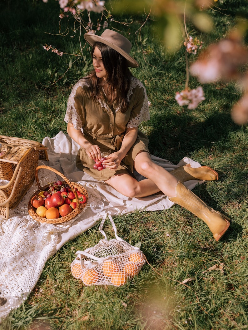 Puede incluir: Una escena de picnic con una mujer con un vestido y sombrero marrones, sentada en una manta en una zona de c&eacute;sped. Una cesta de mimbre llena de fruta, una bolsa de red blanca con naranjas y una cesta tejida est&aacute;n cerca. La mujer sostiene uvas.