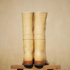 May include: A pair of tall, light tan leather boots with decorative stitching. The boots are displayed on a small wooden stool against a neutral background. The boots have a classic design, with a rounded toe and a stacked heel.