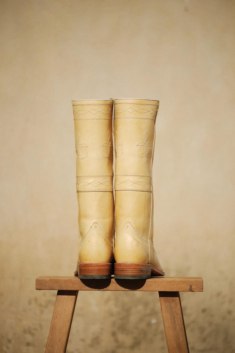 May include: A pair of tall, light tan leather boots with decorative stitching. The boots are displayed on a small wooden stool against a neutral background. The boots have a classic design, with a rounded toe and a stacked heel.