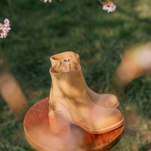 May include: Tan leather ankle boots with a block heel and a side zip. The boots are sitting on a wooden stool. The brand name "SENDRA" is visible on the side of the boot.