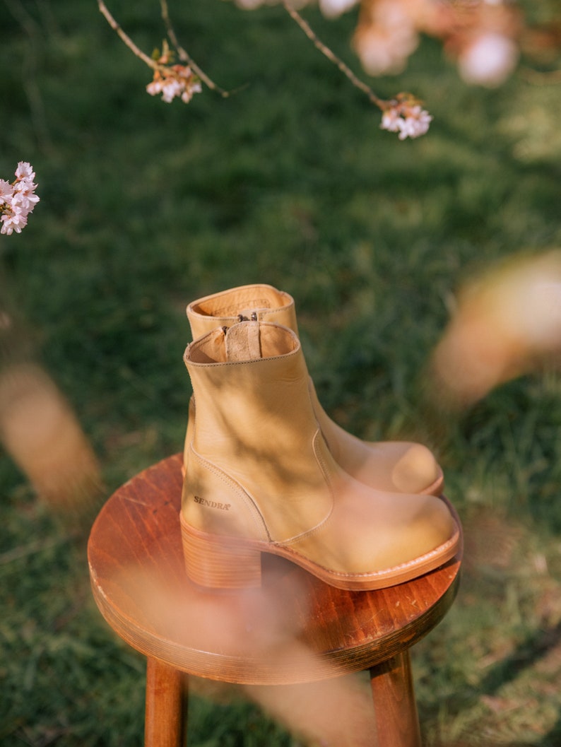 May include: Tan leather ankle boots with a block heel and a side zip. The boots are sitting on a wooden stool. The brand name "SENDRA" is visible on the side of the boot.
