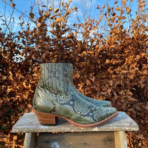 Könnte beinhalten: Grüne Schlangenleder-Cowbo Stiefel mit spitzer Zehenkappe und niedrigem Absatz. Die Stiefel stehen auf einem Holzhocker vor einem Hintergrund aus braunen Blättern.