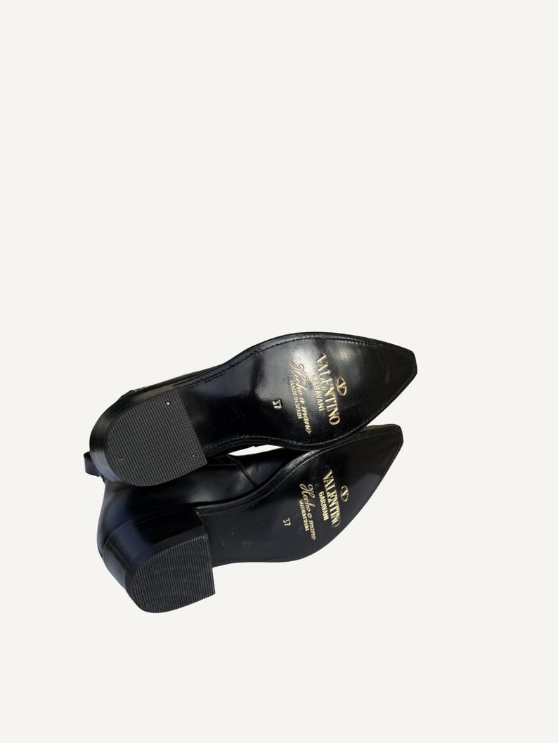 May include: Pair of black leather shoes with a pointed toe and a low block heel. The soles are visible, displaying the brand name "Valentino" in gold lettering. The shoes are size 37.