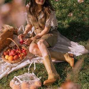 Puede incluir: Una escena de picnic con una mujer con sombrero y vestido, sentada en una manta en una zona de c&eacute;sped. Una cesta de mimbre llena de fruta, una bolsa de red con naranjas y botas vaqueras color canela son visibles.
