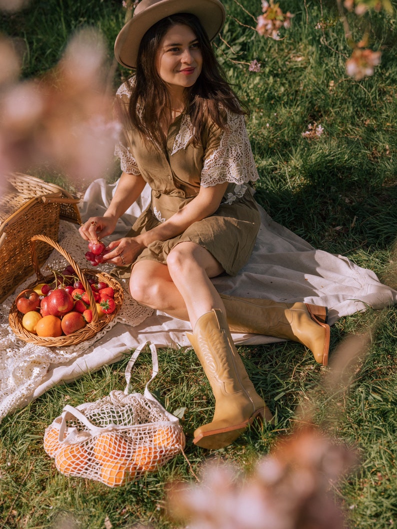 Puede incluir: Una escena de picnic con una mujer con sombrero y vestido, sentada en una manta en una zona de c&eacute;sped. Una cesta de mimbre llena de fruta, una bolsa de red con naranjas y botas vaqueras color canela son visibles.
