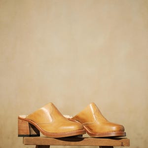May include: A pair of tan leather clogs with a square toe and a block heel. The clogs are displayed on a small wooden stool against a neutral background. The shoes have a Western-inspired design.