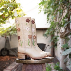 May include: A pair of beige leather boots with a floral design. The boots have a block heel and are sitting on a wooden stool.