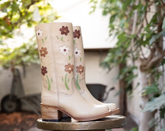May include: A pair of beige leather boots with a floral design. The boots have a block heel and are sitting on a wooden stool.
