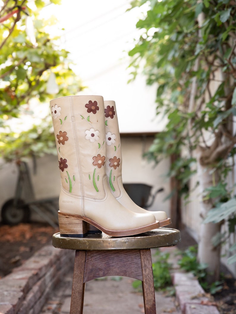 May include: A pair of beige leather boots with a floral design. The boots have a block heel and are sitting on a wooden stool.