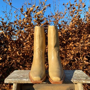 May include: A pair of tan leather cowboy boots with a pointed toe and a square heel. The boots are sitting on a wooden stool in front of a background of brown leaves.