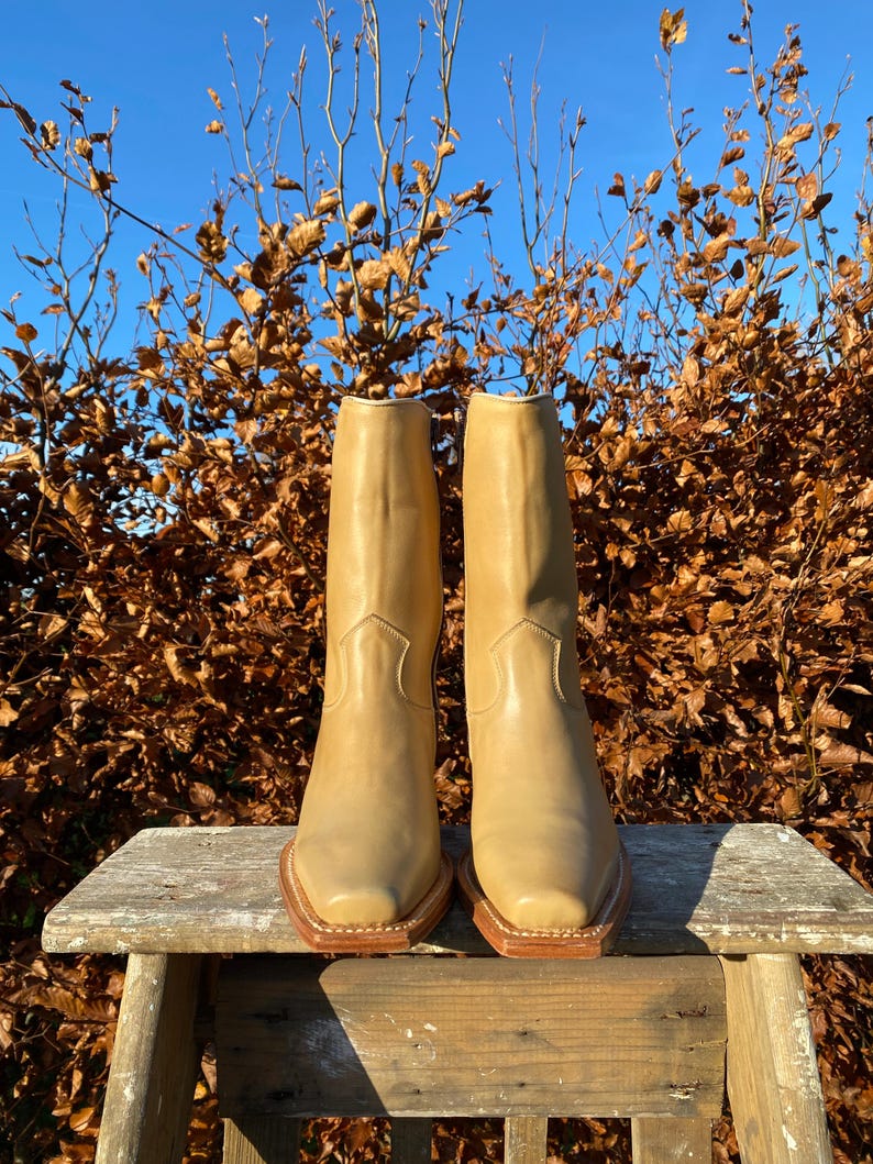 May include: A pair of tan leather cowboy boots with a pointed toe and a square heel. The boots are sitting on a wooden stool in front of a background of brown leaves.