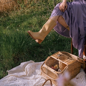 May include: A light tan leather knee-high boot with a block heel. The boot is worn with a lavender dress. A wicker picnic basket and a basket of fruit sit on a white lace blanket in a grassy outdoor setting.