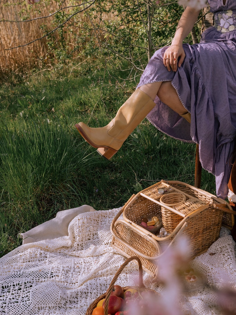 May include: A light tan leather knee-high boot with a block heel. The boot is worn with a lavender dress. A wicker picnic basket and a basket of fruit sit on a white lace blanket in a grassy outdoor setting.
