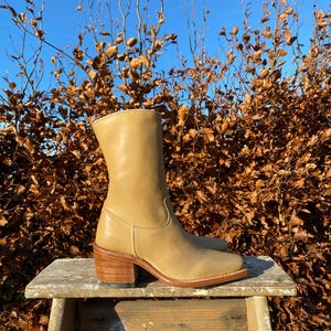 May include: Tan leather cowboy boots with a stacked heel. The boots are sitting on a wooden stool against a backdrop of brown leaves.