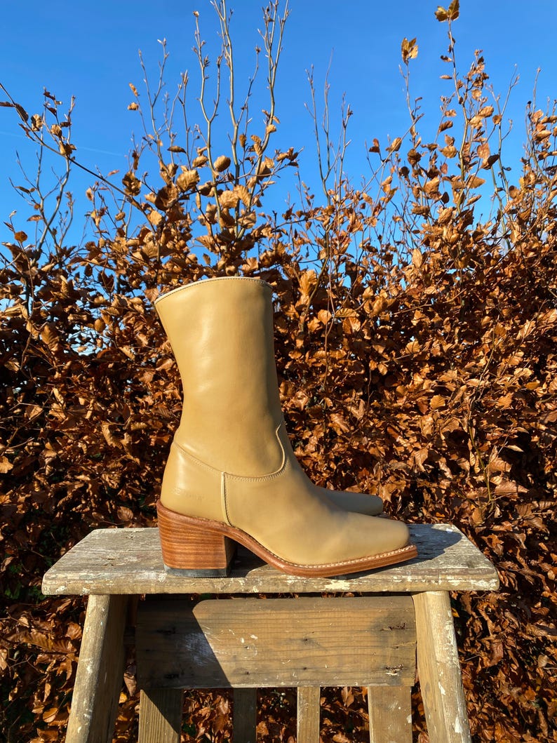May include: Tan leather cowboy boots with a stacked heel. The boots are sitting on a wooden stool against a backdrop of brown leaves.