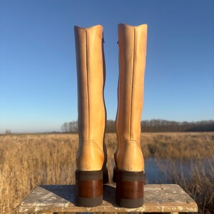 May include: A pair of tall, light brown leather boots with a chunky heel. The boots have a zipper on the inside of each boot and are sitting on a wooden stool.